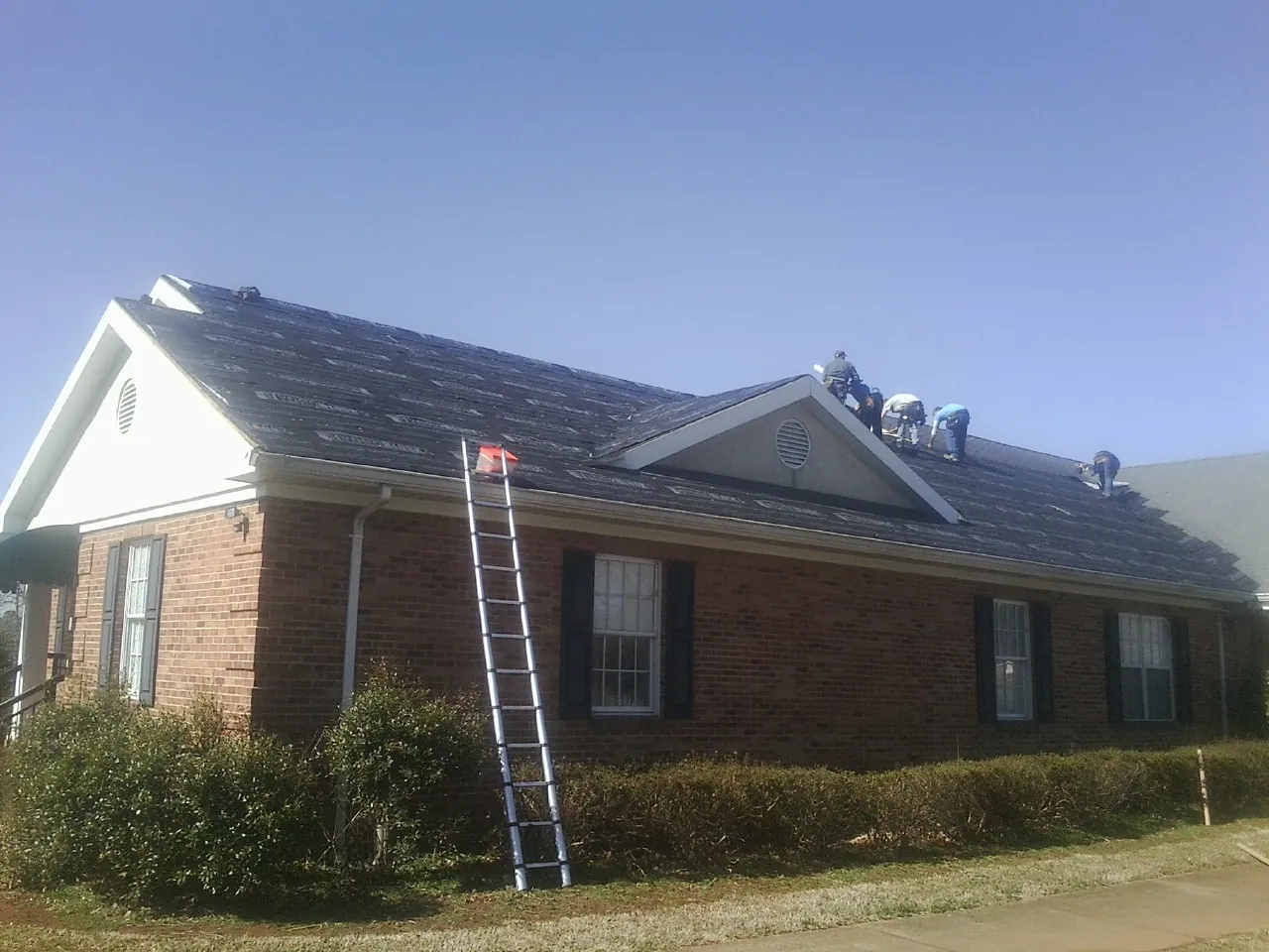 Crew tearing shingles off a brick ranch roof with a ladder set — Emergency Roof Repair work by our crew in New Lisbon